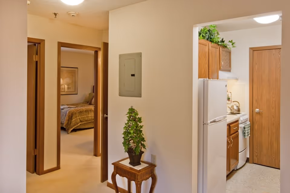 Interior view of a senior living facility showing a small hallway with a wooden table holding a potted plant. To the left, there is a bedroom with a bed covered in a brown blanket. To the right, there is a kitchen area with wooden cabinets, a white refrigerator, and a stove with a kettle on it.