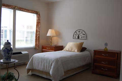 A simple, neatly arranged bedroom with a single bed covered in a white bedspread and a beige pillow. On either side of the bed are wooden nightstands, one with a brass lamp and the other with a small decorative item. A window with floral curtains allows natural light into the room, and a decorative metal wall piece hangs above the bed.