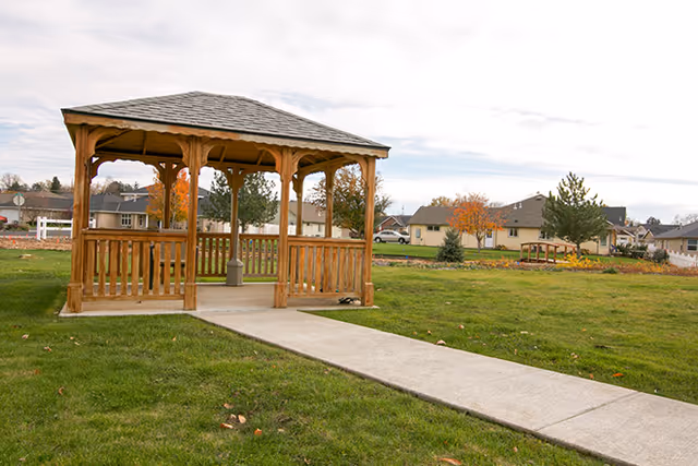 Wooden gazebo on a grassy lawn with a concrete path leading to it and houses in the background.