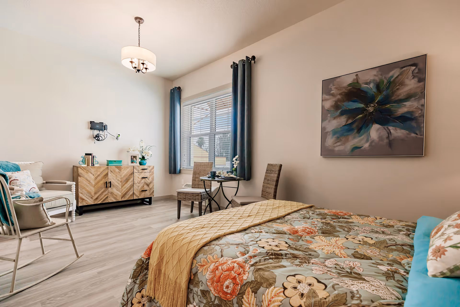 A bright and cozy bedroom featuring a bed with floral bedding and a mustard yellow throw blanket. The room includes a wooden sideboard with decorative items, a small round table with two chairs near a window with blue curtains, a rocking chair, and a large floral painting on the wall.