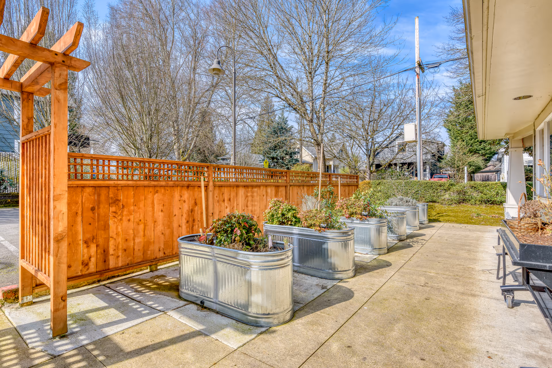 Outdoor patio area with a row of metal planter boxes containing plants, a wooden fence with lattice top, leafless trees, and a building with a covered walkway on the right side under a clear blue sky.