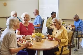 A group of elderly people sitting around a round table in a dining area, enjoying food and drinks while engaging in conversation. The room has a bright window and simple decor.