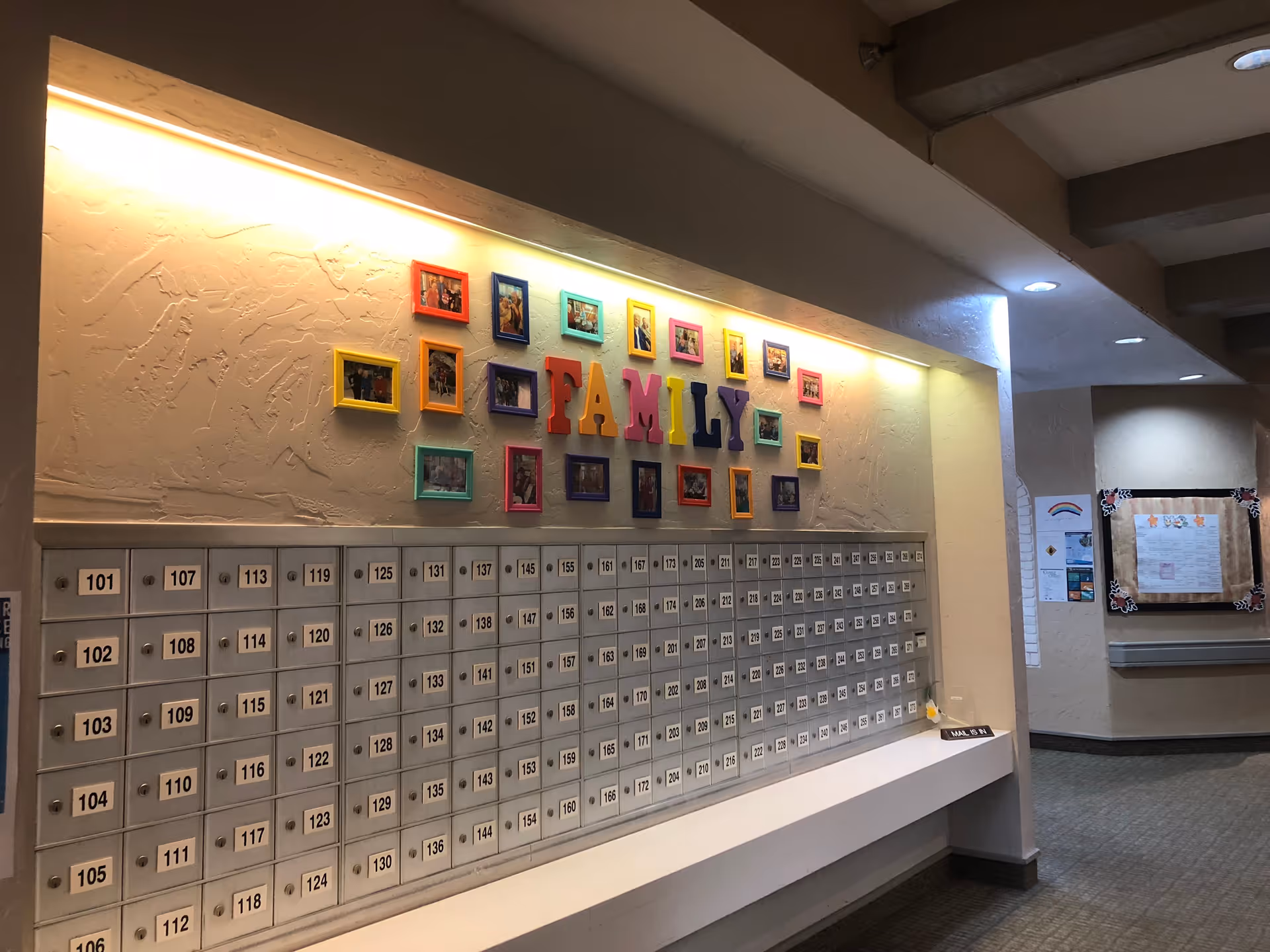 A wall with multiple mailboxes numbered in rows, above which is a decorative display with colorful picture frames and large letters spelling 'FAMILY'. The area is well-lit with overhead lighting and has a textured beige wall. To the right, there is a hallway with bulletin boards and informational posters.