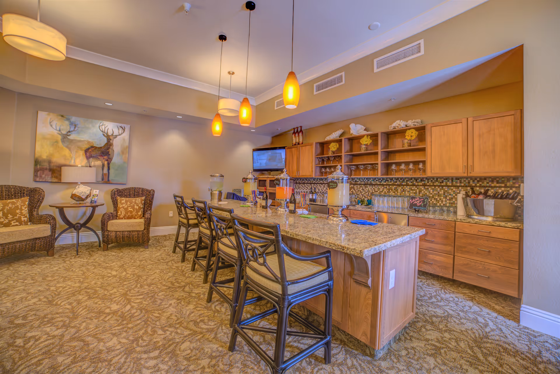 A cozy common area with a long granite countertop island surrounded by six bar stools. The countertop has beverage dispensers and glasses. Behind the island, there are wooden cabinets and shelves with decorative items and glassware. To the left, there are two wicker armchairs with cushions and a small round table between them. The walls are painted beige, and there is a painting of two deer hanging on the wall. The room is warmly lit with pendant lights and ceiling fixtures.