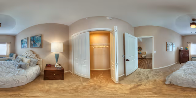 Panoramic view of a bedroom with two beds on opposite sides, each with white bedding and pillows. Between the beds is a nightstand with a lamp and an alarm clock. The room has beige walls and carpeted floor. There is an open closet with hangers in the center and an open door leading to another room with a wooden floor and furniture.