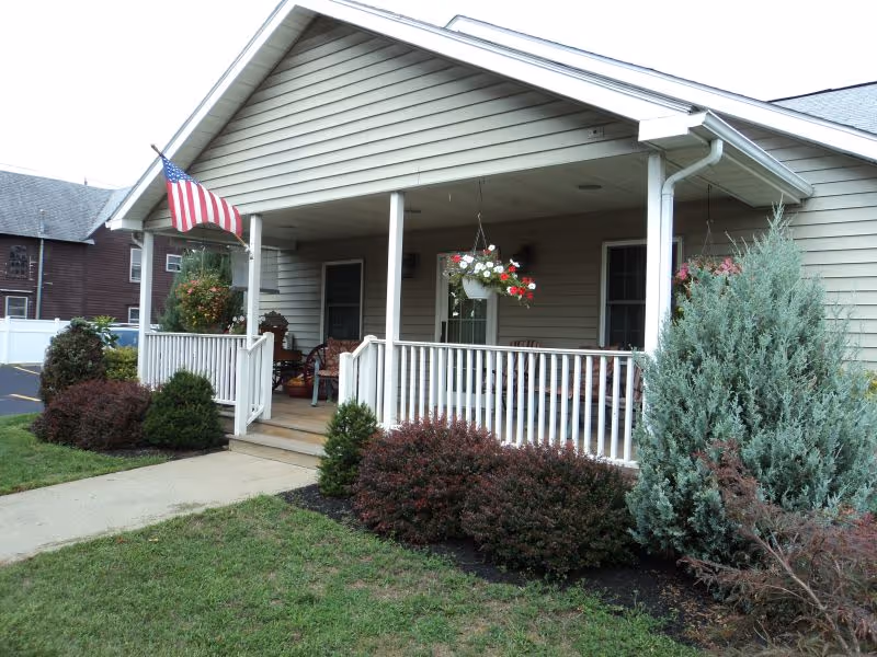 Covered front porch with white railing, hanging flower baskets, an American flag, and landscaped shrubs.