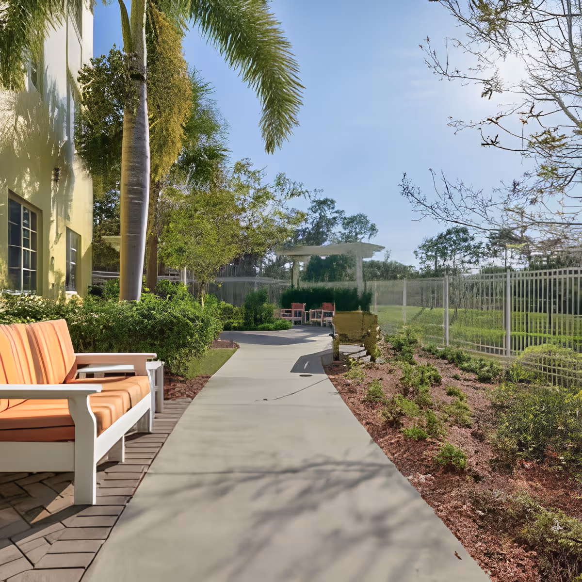 A sunny outdoor walkway at a senior living facility lined with orange-cushioned benches, palm trees, shrubs, and a fence.