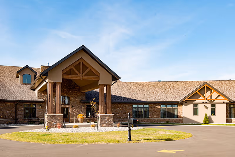 Exterior view of a single-story senior living facility building with a covered entrance supported by wooden pillars, stone and beige walls, and a shingled roof under a clear blue sky.