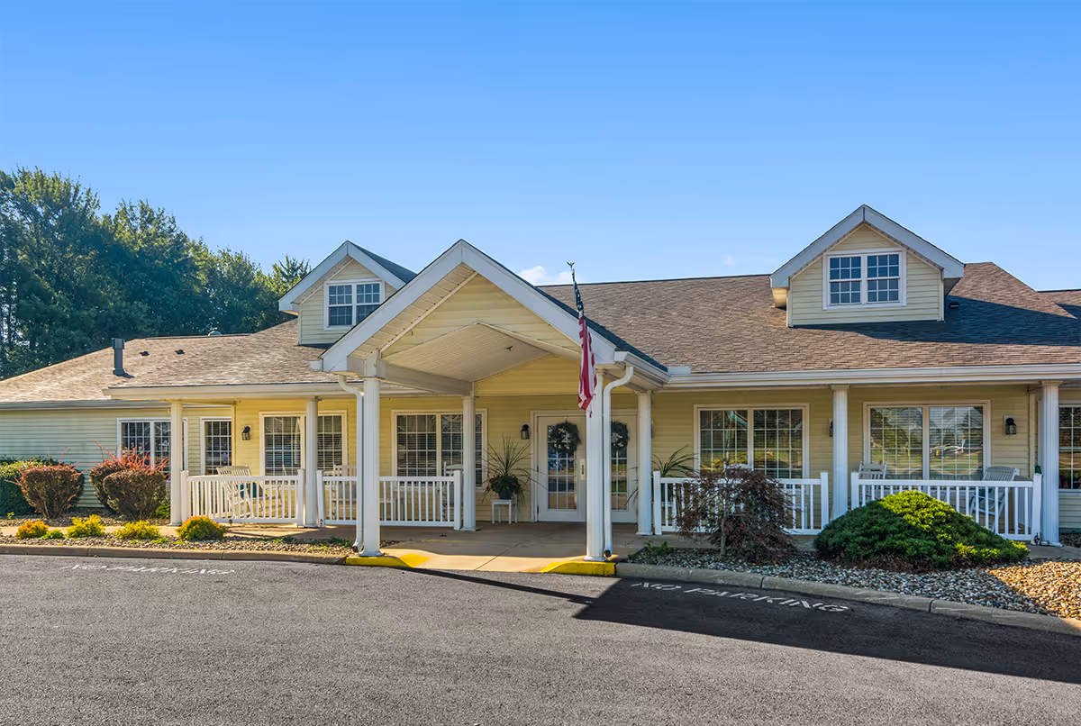 Exterior front view of a single-story building with a covered entrance supported by white columns. The building has a beige facade with white trim, multiple windows, and two dormer windows on the roof. There are shrubs and small plants along the front, and an American flag is displayed near the entrance. The sky is clear and blue.