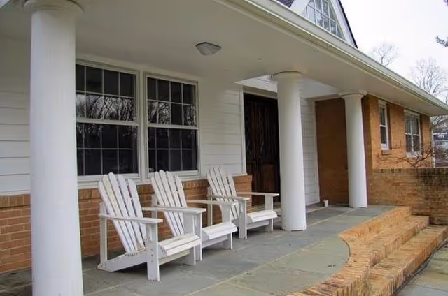 Front porch of a building with three white wooden Adirondack chairs lined up against the wall, large white columns supporting the roof, and brick steps leading up to the entrance.