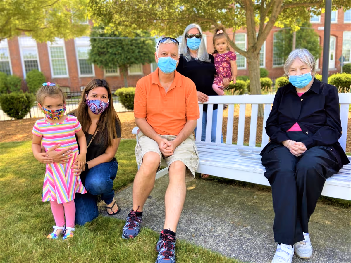A group of six people, including two young children, two middle-aged adults, and two elderly adults, are gathered outdoors in a garden area. They are all wearing face masks. One elderly woman is sitting on a white bench, a man in an orange shirt and beige shorts is sitting next to her, a woman with long gray hair is standing behind the bench holding a young girl, and another woman is kneeling on the grass next to a young girl in a colorful striped dress. Trees and a brick building with large windows are visible in the background.
