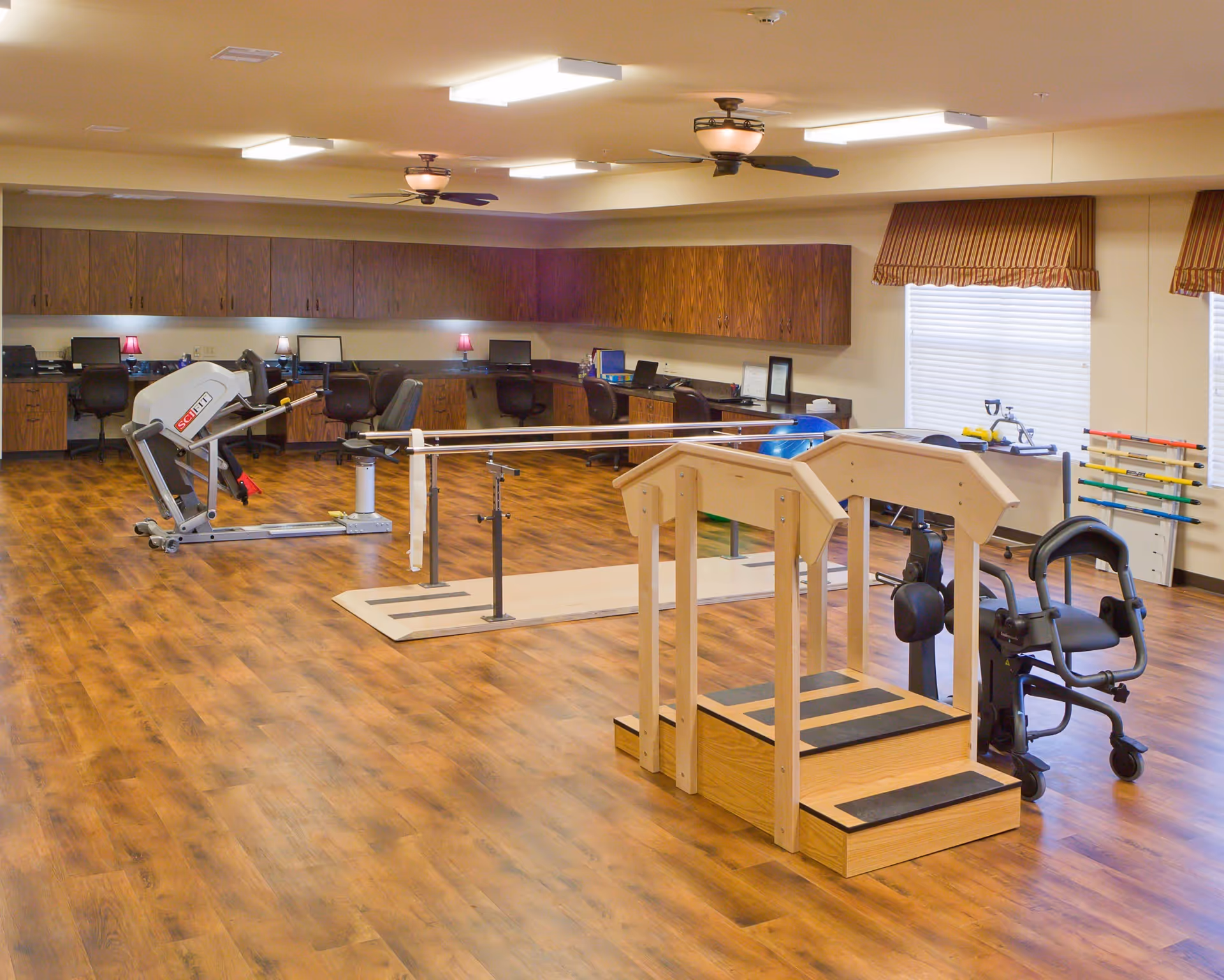 A spacious rehabilitation or physical therapy room with wooden flooring, parallel bars for walking exercises, a wooden step platform with handrails, exercise equipment, and a row of desks with chairs and computers along the back wall. The room is well-lit with ceiling lights and fans, and has windows with striped valances.