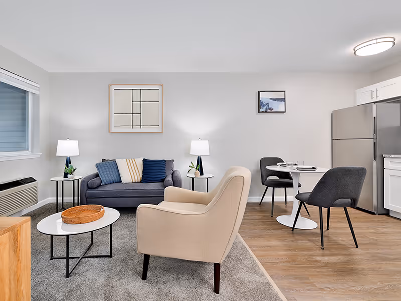 A modern senior living community interior featuring a cozy living room with a gray sofa adorned with blue and beige pillows, a beige armchair, two side tables with lamps, and a white oval coffee table with a wicker tray. Adjacent to the living area is a small dining space with a round white table set for two and two dark gray chairs. The kitchen area includes white cabinetry and a stainless steel refrigerator. The room has light gray walls, wood flooring, and a large window with blinds.