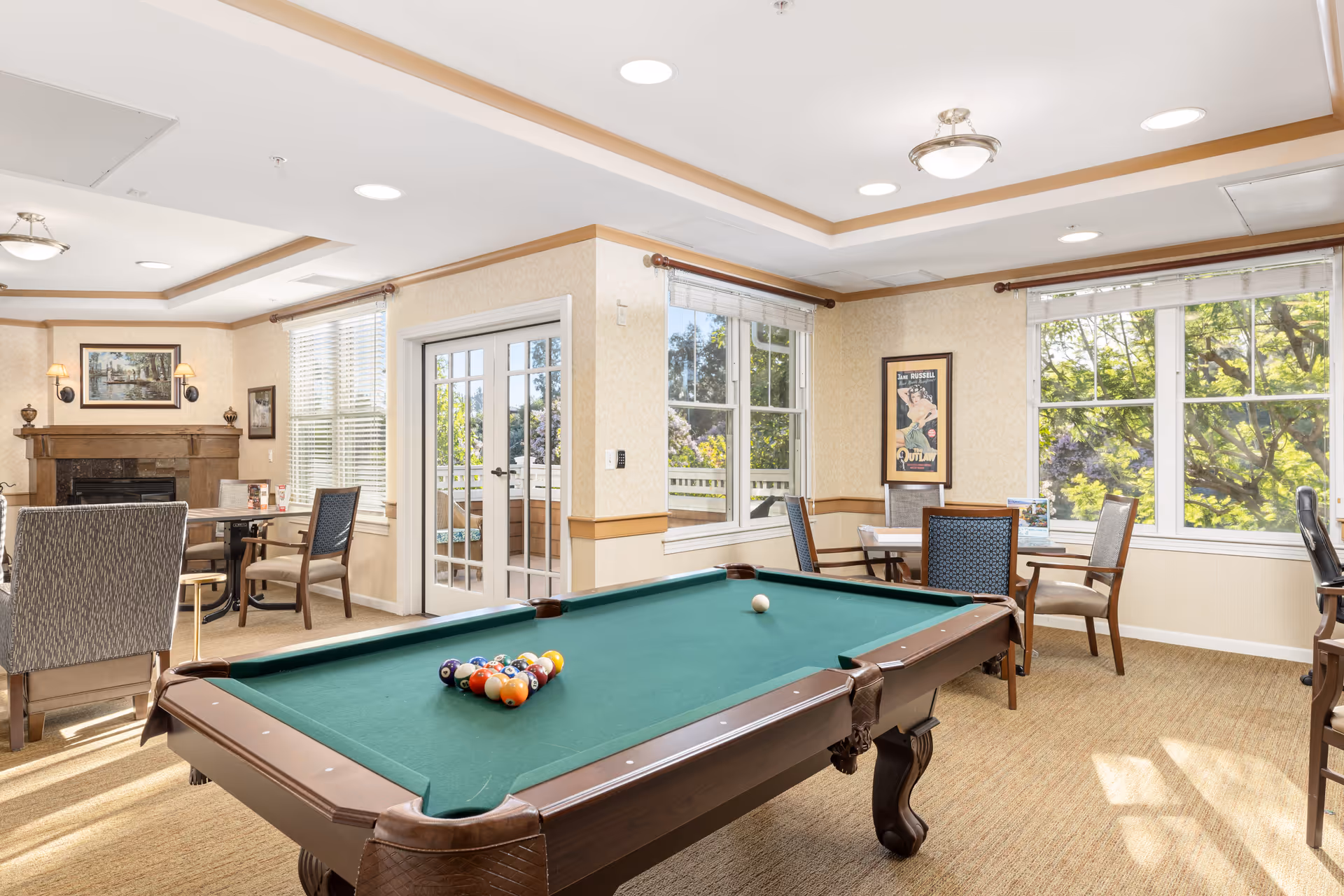 A bright and spacious common room with a pool table in the center, surrounded by several chairs and tables. The room has large windows letting in natural light, a fireplace with artwork above it, and a door leading outside to a patio area. The decor is warm and inviting with beige walls and carpeted floors.