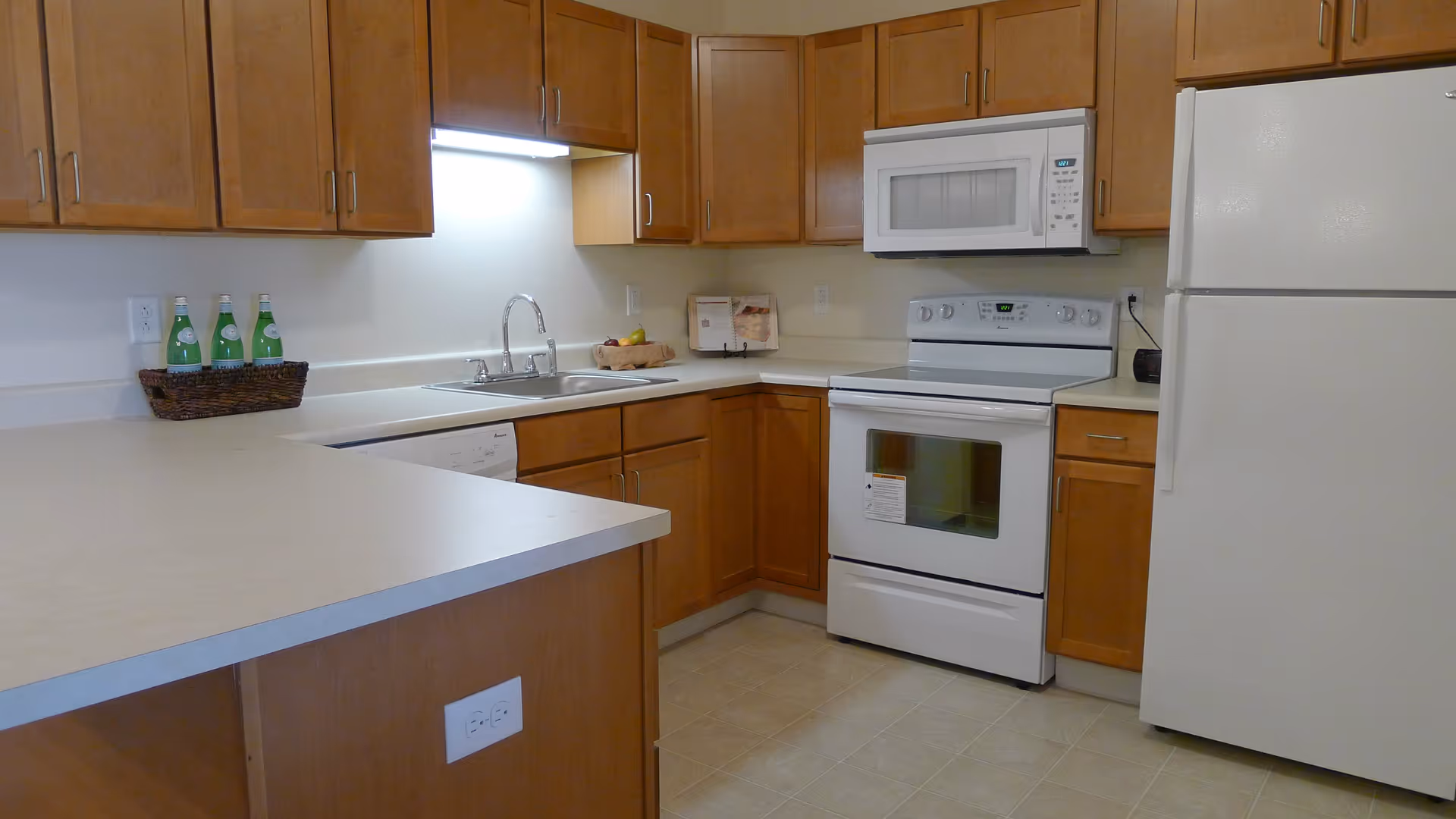 A clean kitchen with wooden cabinets, a white refrigerator, white stove with oven, white microwave, and a double sink. There is a basket with bottles on the countertop and a small fruit basket near the corner.