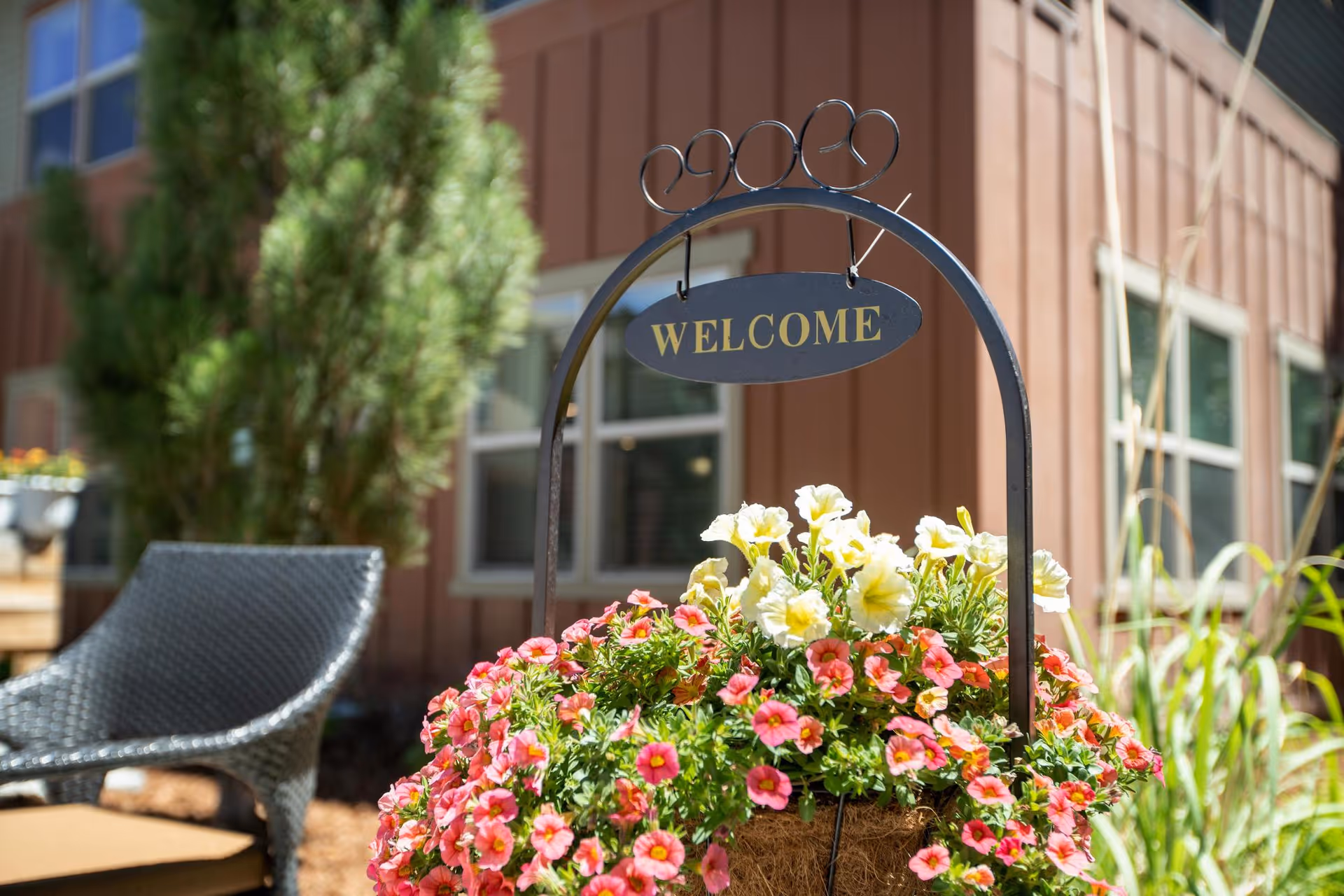 A hanging planter of pink and white flowers under a metal 'WELCOME' sign in an outdoor courtyard by a building.
