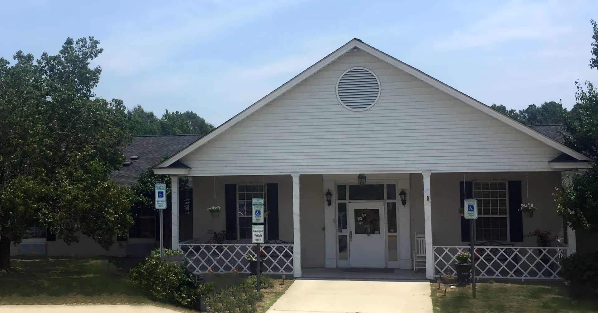 Front exterior view of a single-story building with a white facade, a triangular roof with a circular vent, and a covered porch area with white railings. There are two handicap parking signs in front of the building, surrounded by green trees and shrubs under a partly cloudy sky.