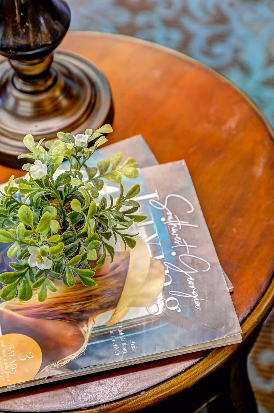 Close-up of a wooden round table with a small green potted plant and two magazines on it. A lamp base is partially visible in the background, and the floor has a patterned carpet.