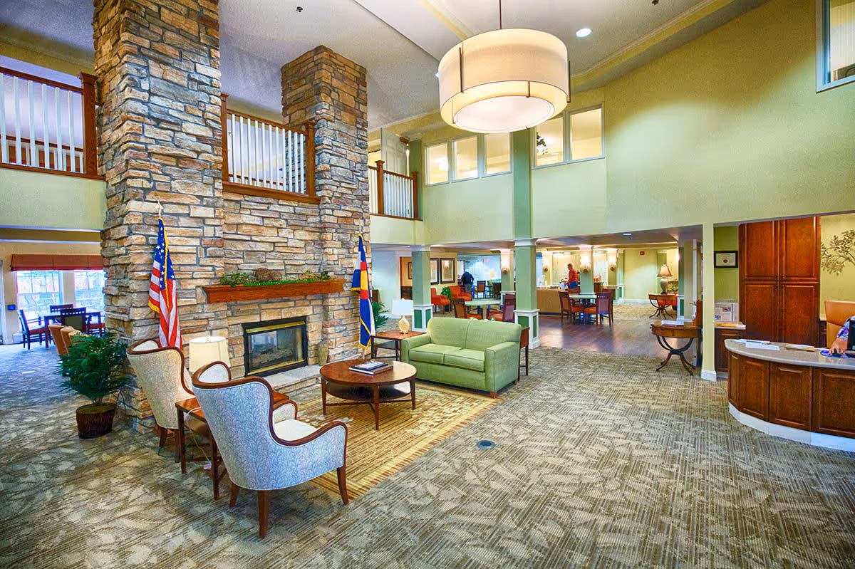 Spacious retirement community common area with a large stone fireplace, green sofa, two upholstered chairs, wooden coffee table, and a large hanging light fixture. The room has carpeted floors, light green walls, and a reception desk on the right side. There are American and Colorado state flags near the fireplace, and a second-floor balcony overlooking the area.