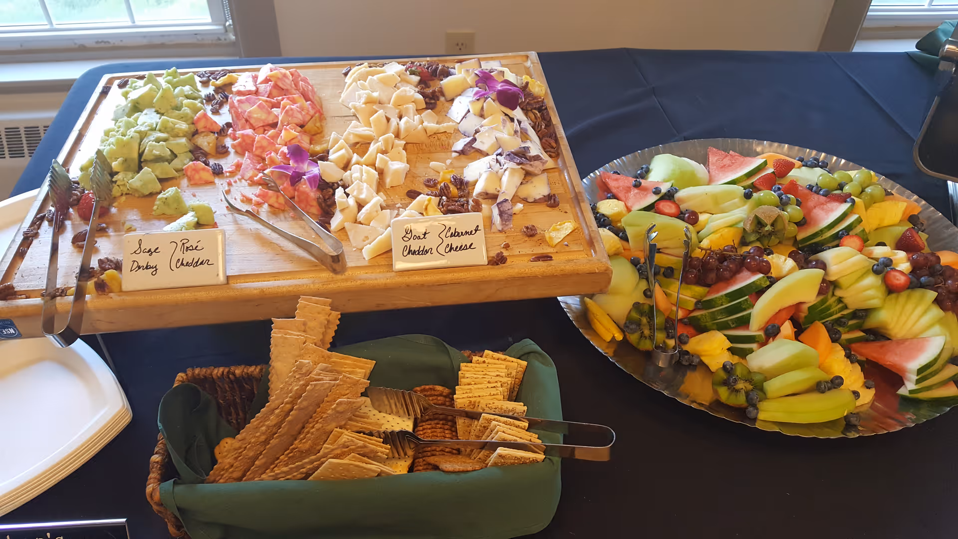 A table set with a wooden board displaying three types of cheese labeled Sage Derby, Rosé Cheddar, and Goat Cheddar Cheese, accompanied by pecans and decorative purple flowers. Next to the cheese board is a large silver platter filled with an assortment of fresh fruit including watermelon, grapes, kiwi, pineapple, strawberries, blueberries, and melon slices. In front of the cheese board is a basket lined with a green cloth containing various types of crackers. The setup is on a black tablecloth near a window.