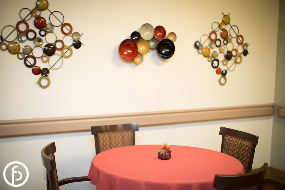 Round table with a red tablecloth and small centerpiece surrounded by chairs beneath decorative circular wall art.