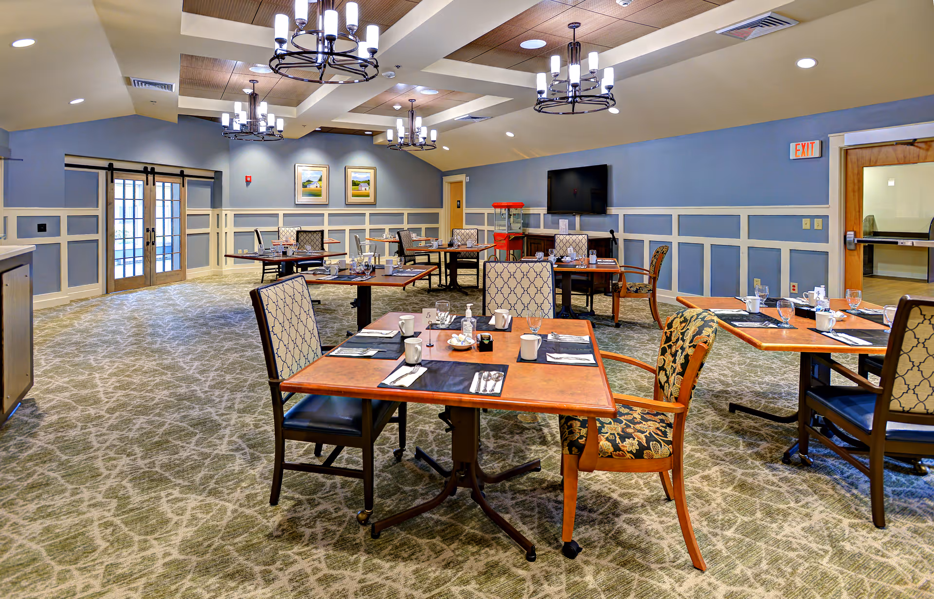 A spacious dining room with several square wooden tables set with cups, glasses, napkins, and utensils. The room has patterned carpet flooring, blue walls with white wainscoting, and multiple chandeliers hanging from the ceiling. There are a few framed pictures on the far wall, a TV mounted on the wall, and double glass doors on the left side.