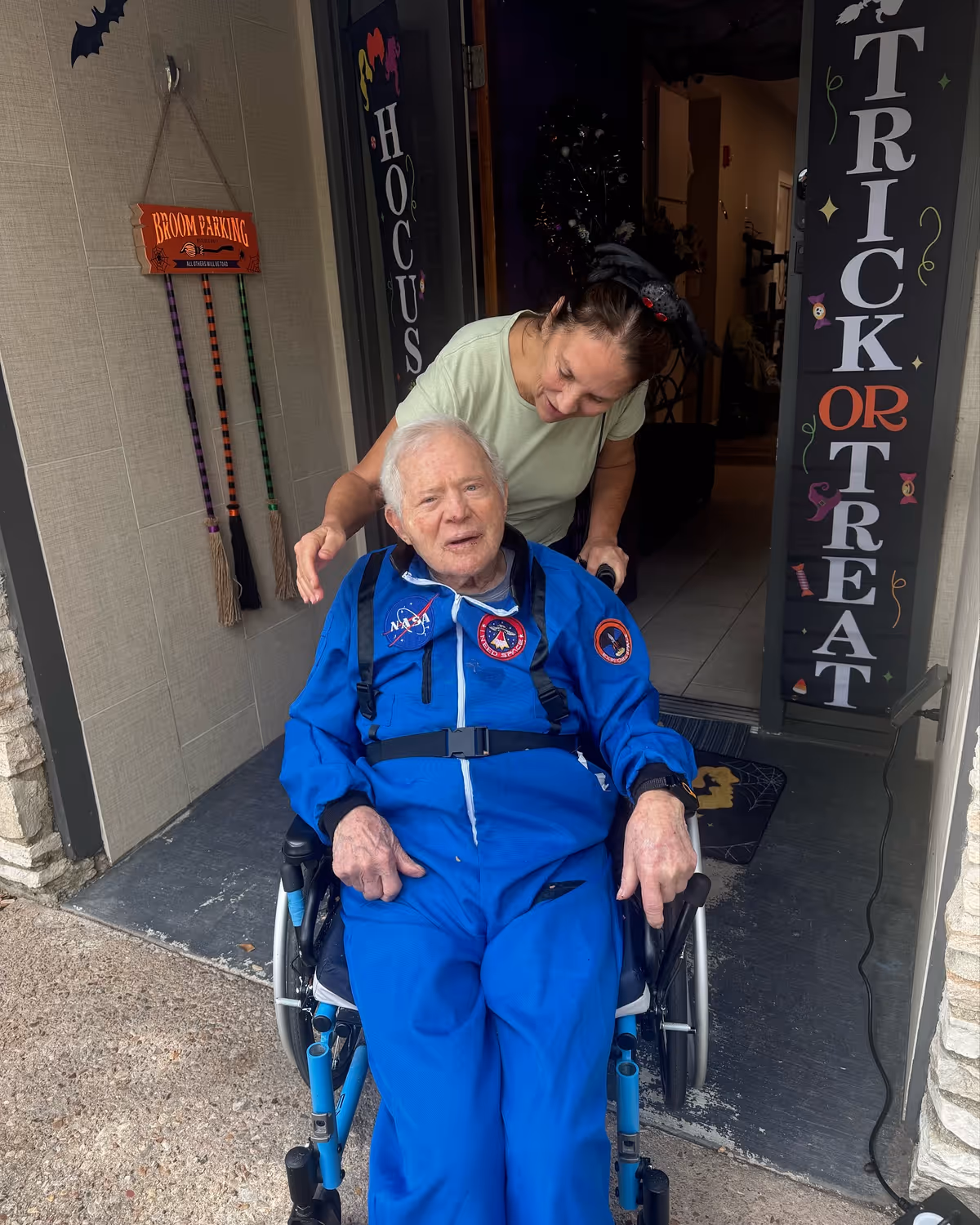 An elderly man in a blue NASA jumpsuit sits in a wheelchair at a decorated entrance while a caregiver leans over him.