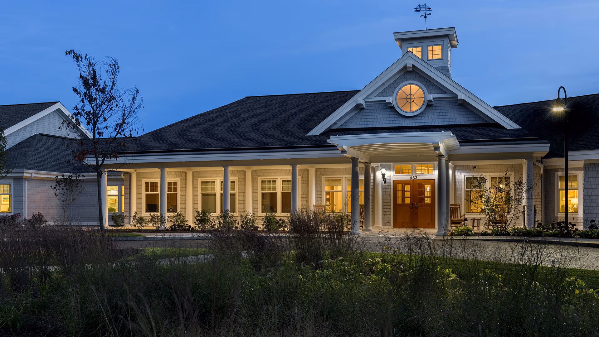 Exterior view of a senior living facility building at dusk with warm lights glowing from the windows. The building features a covered entrance with white columns, a weather vane on the roof, and a landscaped garden area in the foreground.