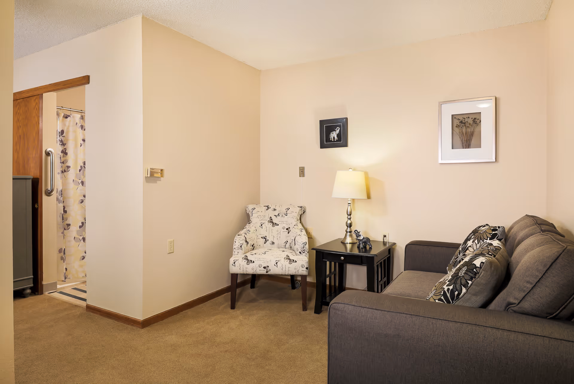 A cozy living area with a gray sofa adorned with patterned cushions, a white armchair with black designs, and a black side table with a lamp and small decorative elephant. The walls are beige with two framed pictures hanging. To the left, there is an open doorway leading to a bathroom with a floral shower curtain.