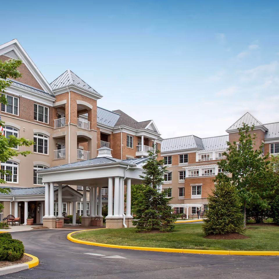 Exterior view of a multi-story senior living facility with brick and beige walls, metal roofs, balconies, and a covered entrance driveway surrounded by green trees and landscaping under a clear blue sky.