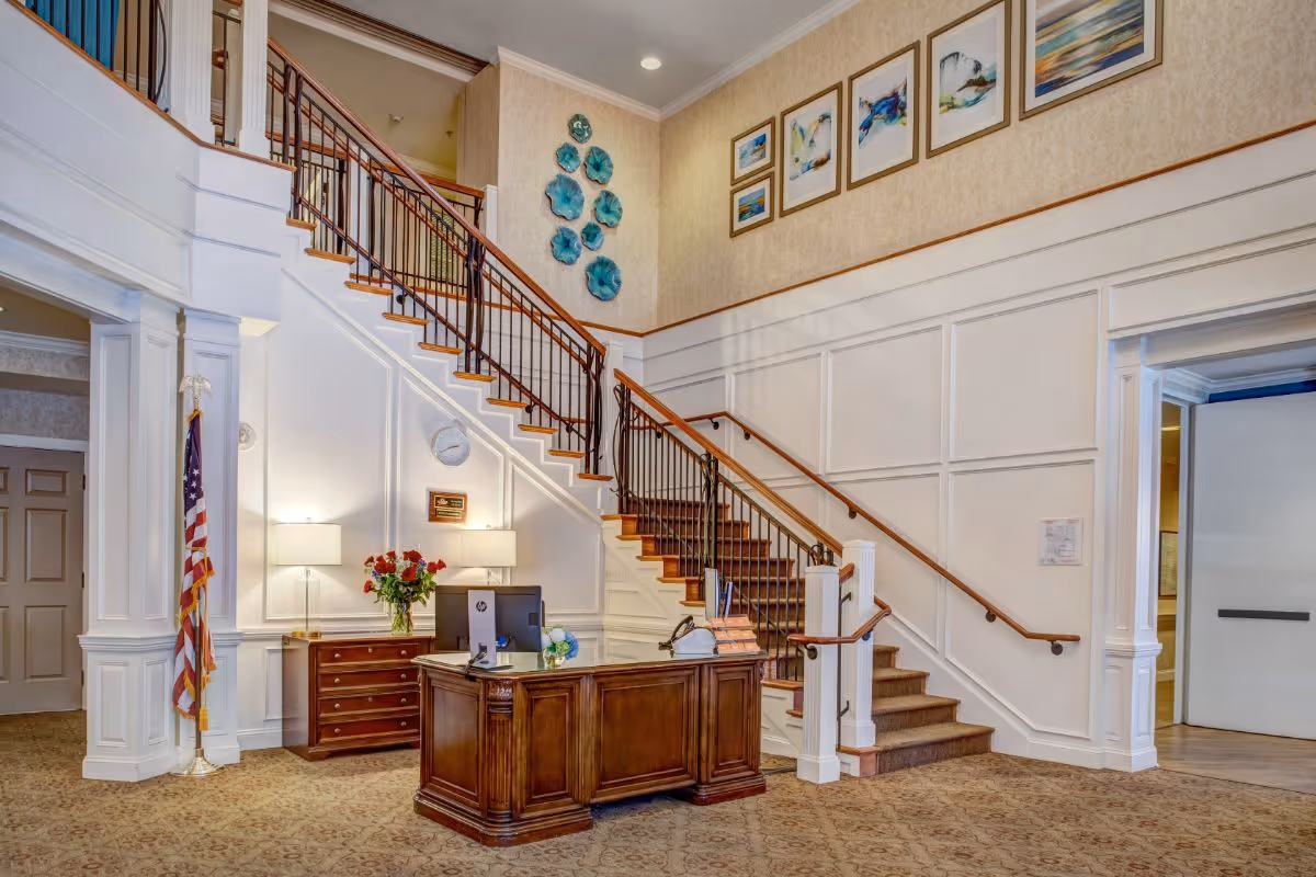 Interior view of a senior living facility lobby with a wooden reception desk, a computer, and a vase of flowers. Behind the desk is a staircase with wooden handrails and black metal balusters leading to an upper floor. The walls are decorated with framed artwork and blue decorative wall pieces. There are two table lamps on a side table next to the desk, and an American flag stands in the corner.