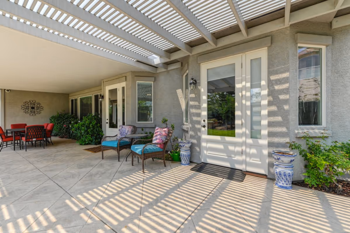 Outdoor patio area with a white pergola casting striped shadows on the tiled floor. There are two wicker chairs with blue cushions and floral pillows, a small round table between them, and a set of double glass doors leading inside. To the left, there is a dining table with red cushioned chairs and some green plants along the wall.