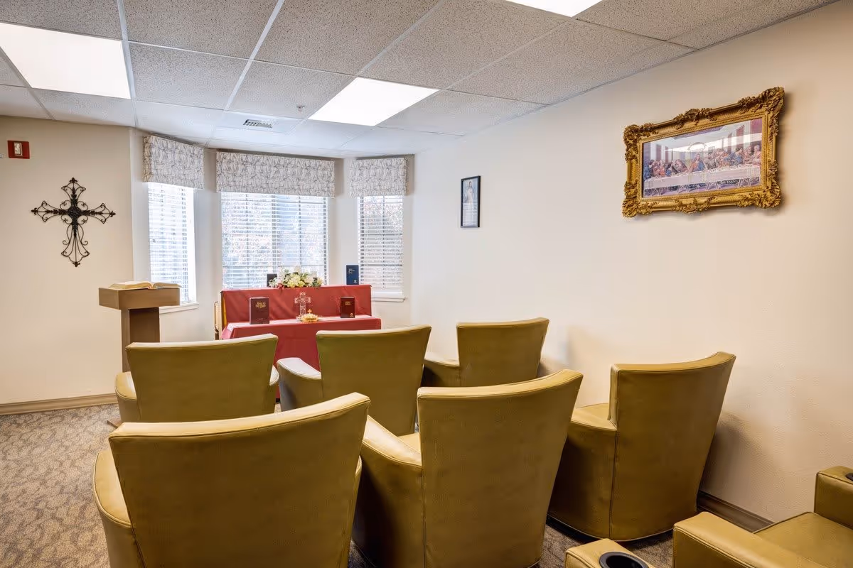 Small chapel room with six green upholstered chairs arranged in two rows facing a small altar covered with a red cloth. The altar has religious books, a cross, and flowers. There is a wooden lectern with an open book on the left side, a decorative cross on the wall, and a framed picture of The Last Supper on the right wall. Three windows with blinds and valances are behind the altar.