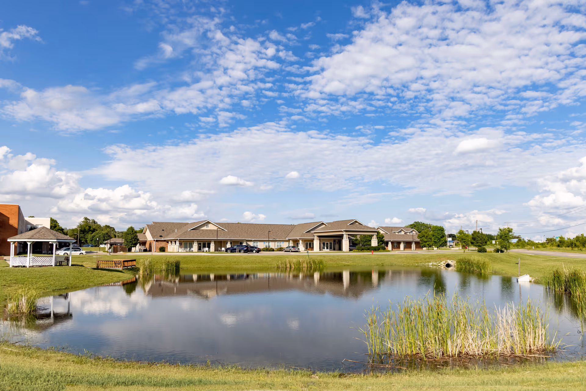 Wide exterior view of Paramount Community Living and Rehab building with a large pond in the foreground, surrounded by grass and some reeds. The sky is partly cloudy with blue patches, and there is a small gazebo and a few parked cars near the building.