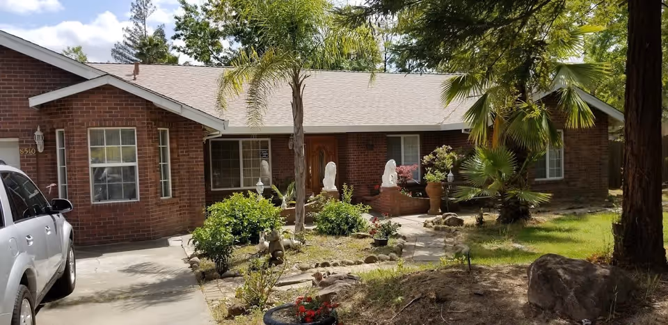 Front exterior view of a single-story brick residential building with a light-colored roof, surrounded by greenery including palm trees, shrubs, and a small garden area. A silver SUV is parked on the driveway to the left, and a stone pathway leads to the wooden front door. The sky is partly cloudy with some blue visible.