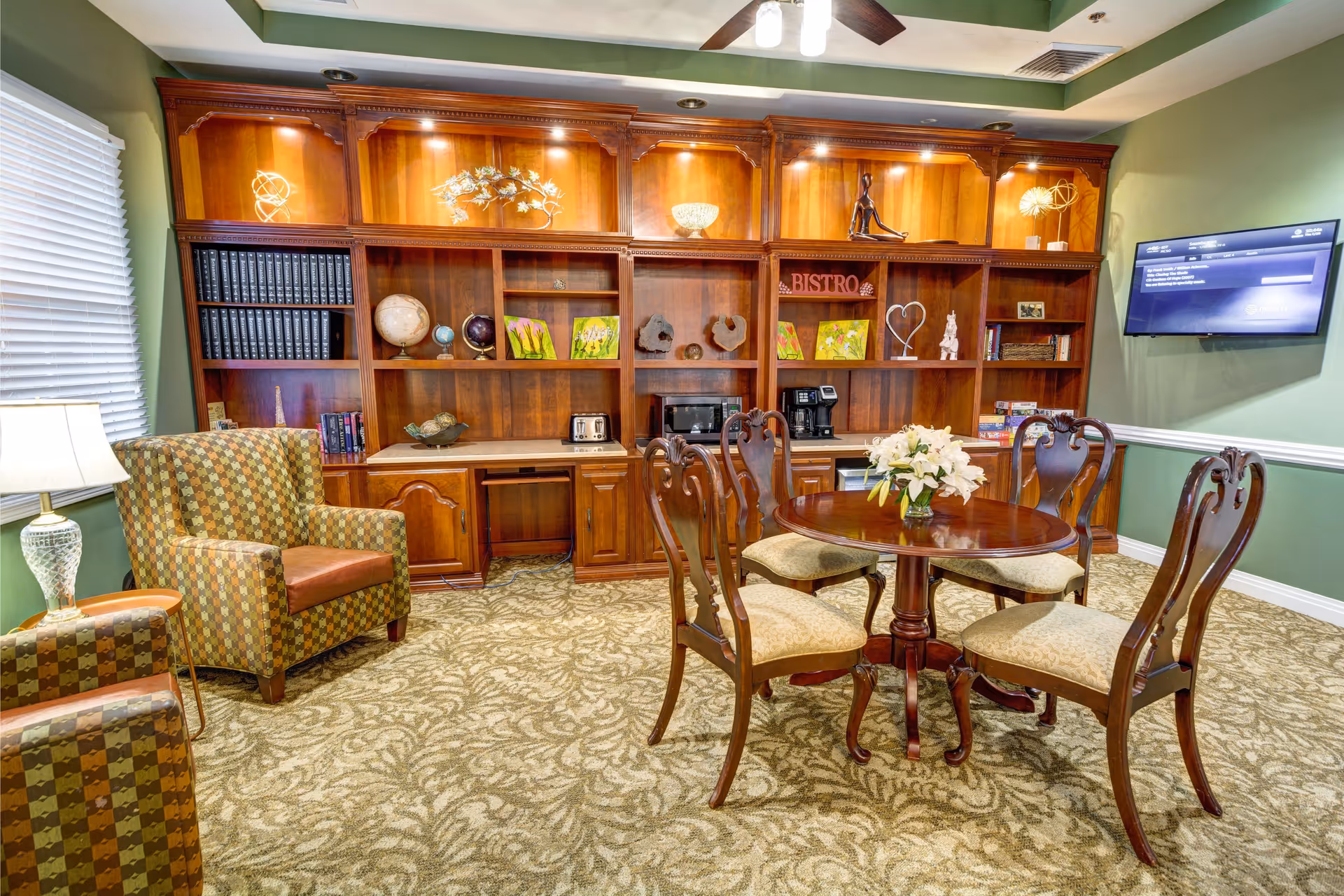 A cozy common area with a round wooden table surrounded by six cushioned chairs. Behind the table is a large wooden built-in shelving unit with decorative items, books, and a microwave. Two patterned armchairs are positioned near a window with blinds and a table lamp. A flat-screen TV is mounted on the green wall to the right.
