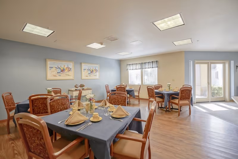 A bright dining room with several tables covered in blue tablecloths, each set with beige napkins, cups, glasses, and silverware. The room has wooden floors, cushioned chairs with patterned upholstery, two framed beach-themed paintings on a blue wall, a window with a valance, and a glass door letting in natural light.