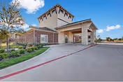 Front entrance and porte-cochère of a brick senior living building with a driveway, landscaped beds, and blue sky.