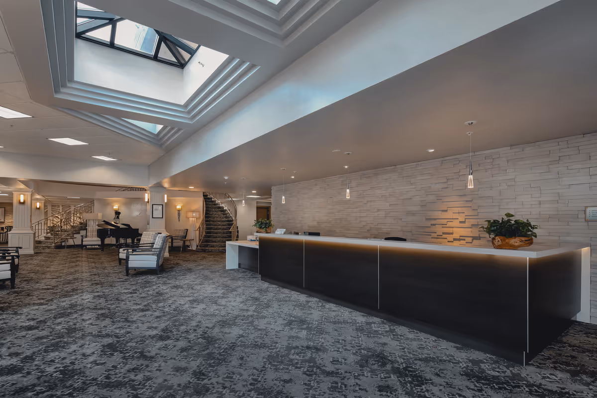 A spacious and modern reception area with a long dark wood front desk illuminated by pendant lights. The floor is covered with patterned carpet, and there are comfortable chairs arranged near a grand piano. A staircase with a decorative railing is visible in the background, and a skylight above allows natural light to brighten the space.