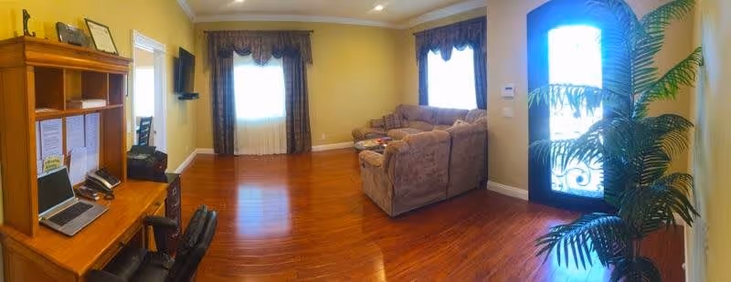 A cozy living room with wooden flooring, a large brown sectional sofa, a wooden desk with a laptop and office supplies, a wall-mounted TV, two windows with curtains, and a decorative plant near a door with frosted glass panels.