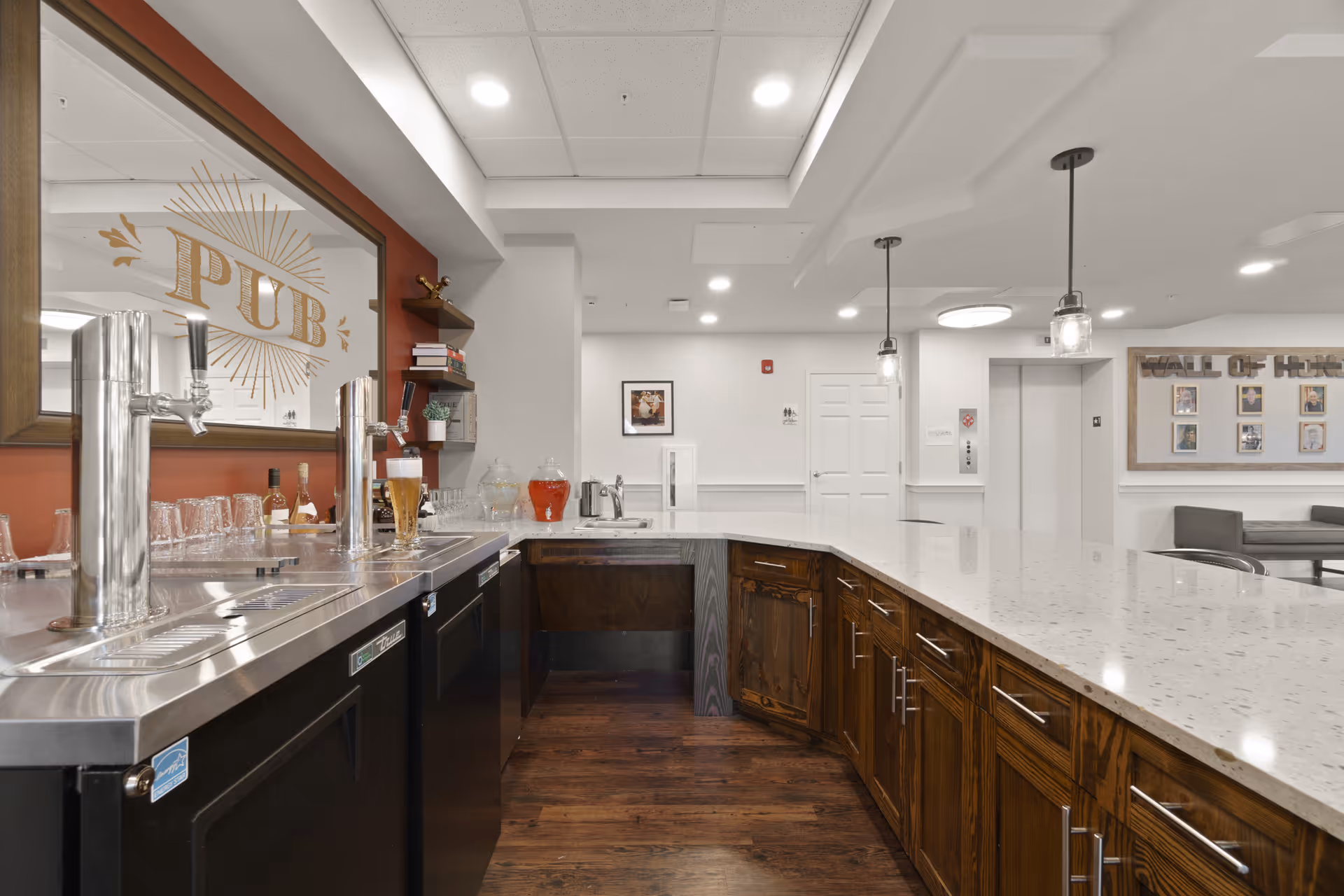 Interior view of a senior living facility's pub area with a long countertop, wooden cabinets, beer taps, glasses, and bottles. The area is well-lit with ceiling lights and pendant lights, and there is a framed mirror with the word 'PUB' on the wall. In the background, there is a hallway with a door, an elevator, and a seating area with a 'Wall of Hope' display.