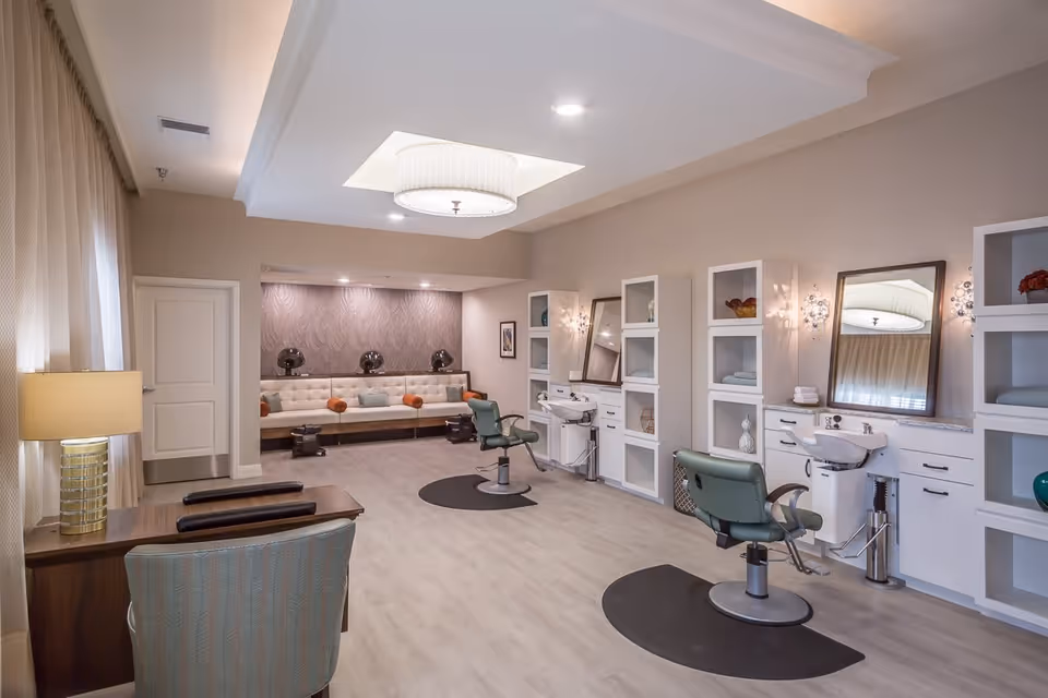 A spacious and well-lit salon area in a senior living facility with two green salon chairs in front of sinks and mirrors, white shelving units with decorative items, a seating area with a long cushioned bench and orange pillows, a desk with a lamp and chair near a window with curtains, and a modern ceiling light fixture.