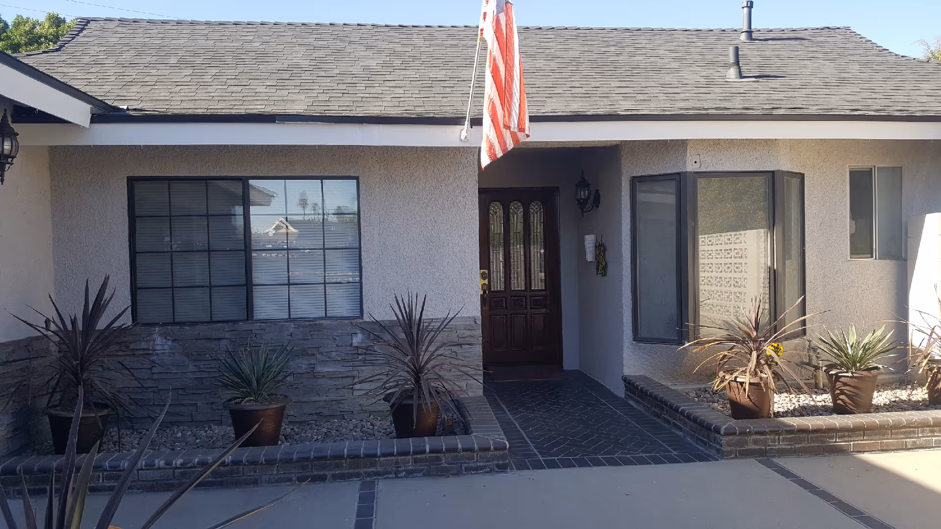 Front exterior view of a single-story building with a dark shingled roof, beige walls, and a wooden front door. There are several potted plants arranged along the front, and an American flag is mounted above the entrance.