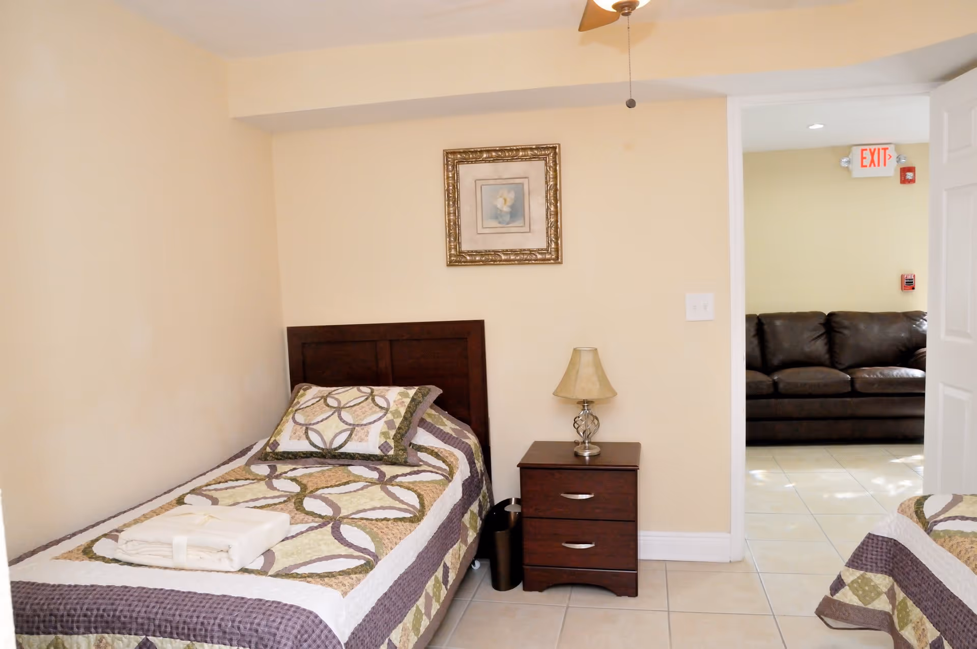 A small bedroom with a single bed covered with a patterned quilt and a folded towel on top. Next to the bed is a wooden nightstand with a lamp. A framed picture hangs on the beige wall above the nightstand. An open door reveals a room with a dark brown leather couch and an exit sign above the door.