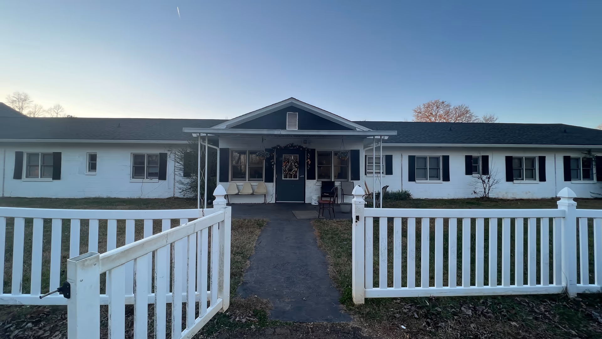 Front exterior view of a single-story white building with black shutters and a dark roof, featuring a covered entrance with a door decorated with a wreath. There is a white picket fence with an open gate leading to the entrance, and some chairs are placed on the porch. The sky is clear and it appears to be late afternoon or early evening.