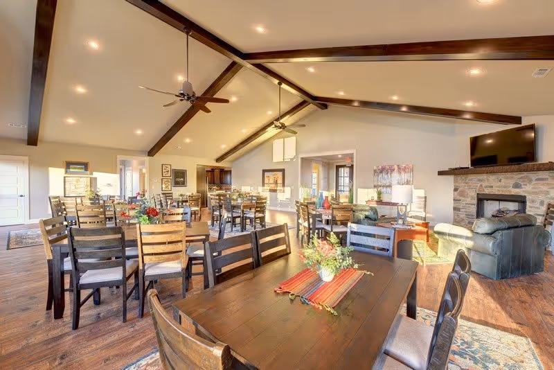 A spacious, well-lit common area with multiple wooden dining tables and chairs arranged neatly. The room features a high ceiling with exposed dark wooden beams and ceiling fans. There is a stone fireplace with a mounted flat-screen TV above it, and a comfortable seating area with leather armchairs near the fireplace. The floor is wooden, and the room is decorated with plants and framed artwork on the walls.