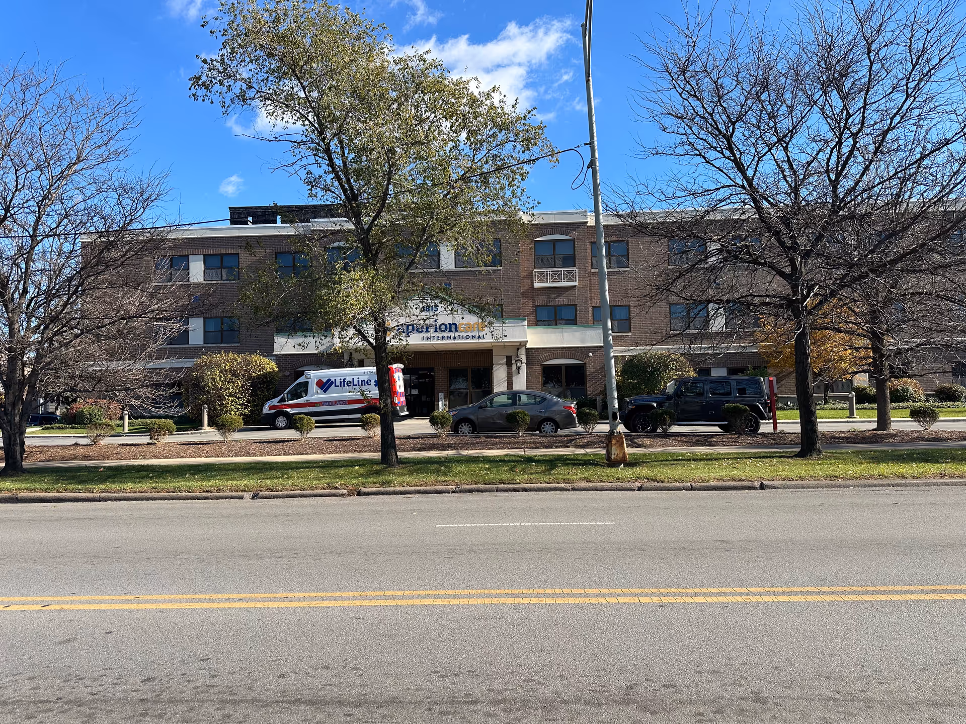 Exterior front view of a three-story brick building with a sign that reads Aperion Care International above the entrance. There are several trees, some with leaves and some bare, lining the sidewalk in front of the building. Three vehicles, including a LifeLine ambulance van, are parked in front of the building. The sky is clear and blue with a few clouds.