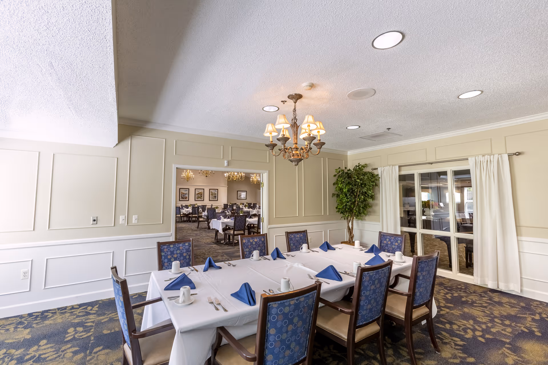 Formal dining room with a long table set with a white tablecloth, blue napkins and multiple chairs beneath a chandelier.