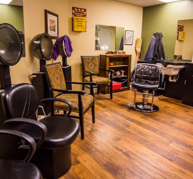 Small salon room with barber chair, hooded hair dryers, seating chairs, mirrors and a shampoo sink on a wood floor.