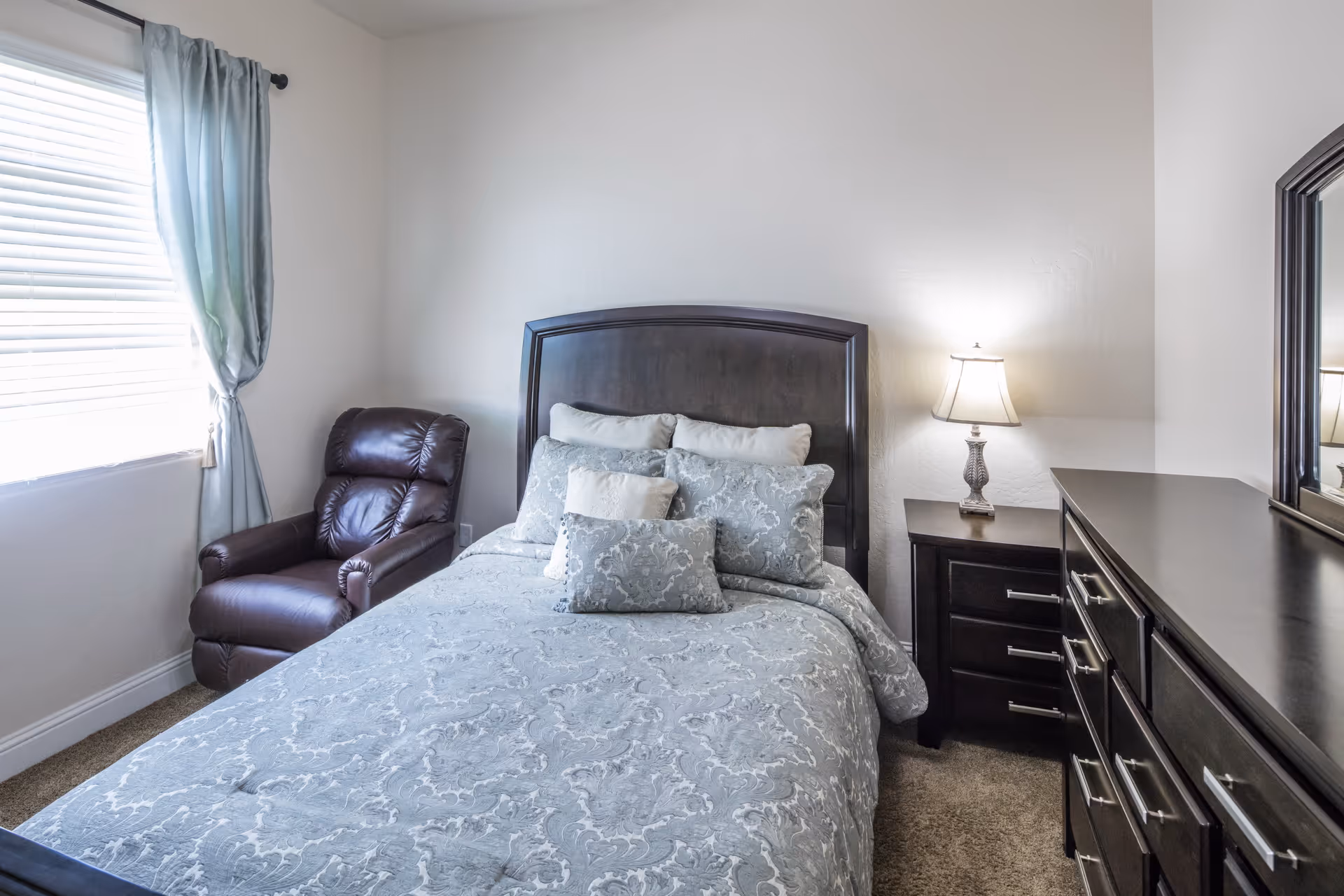 A neatly made bed with patterned bedding and multiple pillows in a bedroom. To the left of the bed is a brown leather recliner chair near a window with light blue curtains. On the right side of the bed is a dark wooden nightstand with a lamp, and next to it is a matching dresser with a large mirror.
